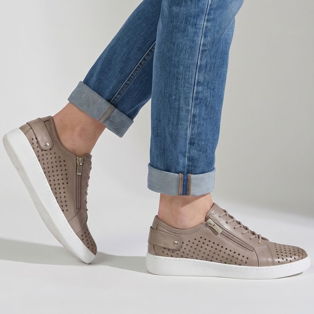 A pair of taupe perforated leather low-top sneakers with white soles, shown on a model wearing blue jeans against a light gray studio background.