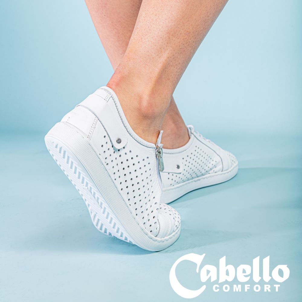 A person wearing a pair of white sneakers with a perforated Turkish leather upper, side zip entry and lace-up adjustment, against a blue background.
