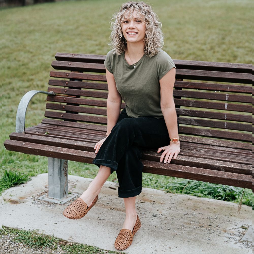 A lady wearing a green t-shirt and black pants, crossing her legs as she is sitting on a park bench wearing a pair of tan laser-cut Cabello leather loafers.