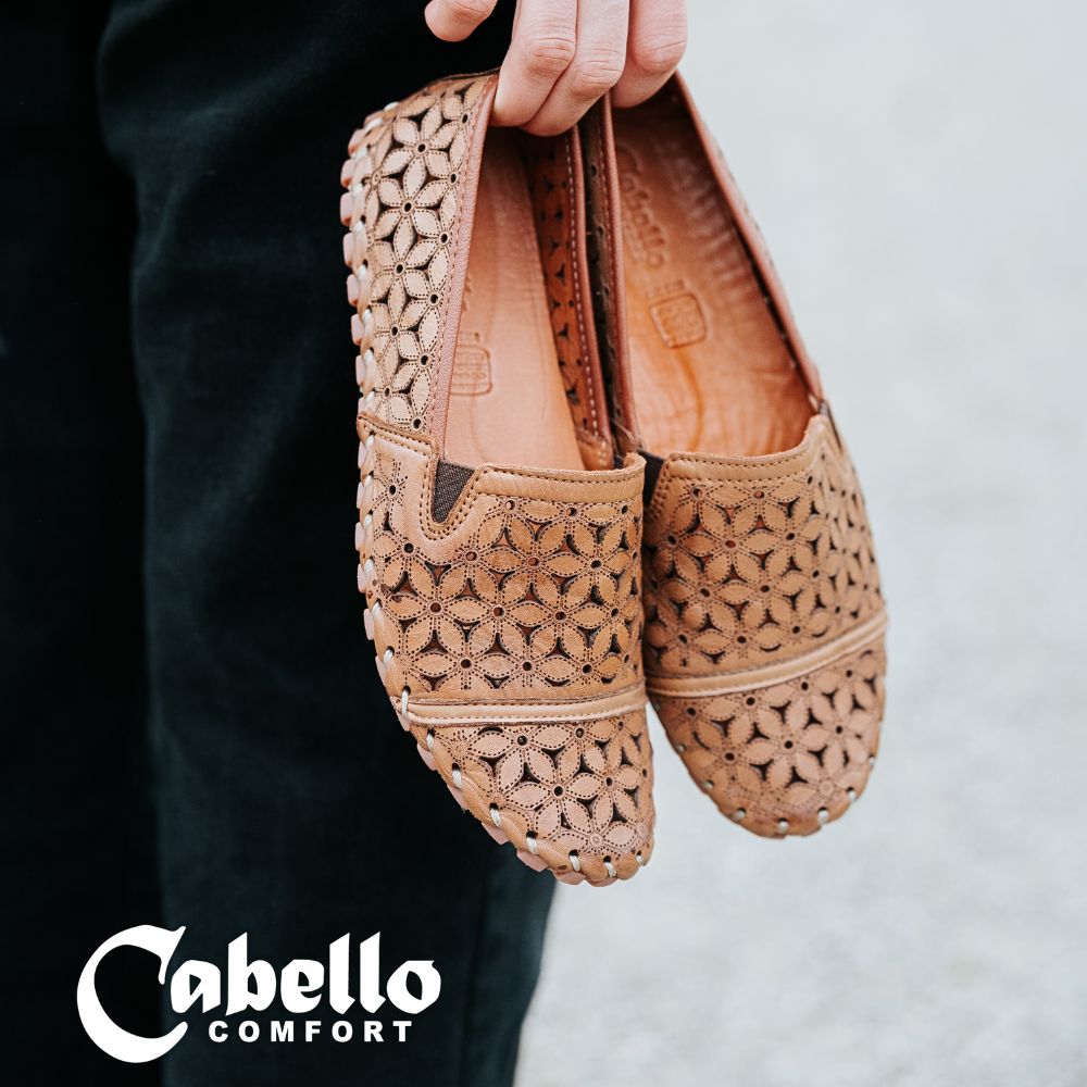 A person wearing black trousers, holding a pair of tan laser-cut Cabello leather loafers by the heels.