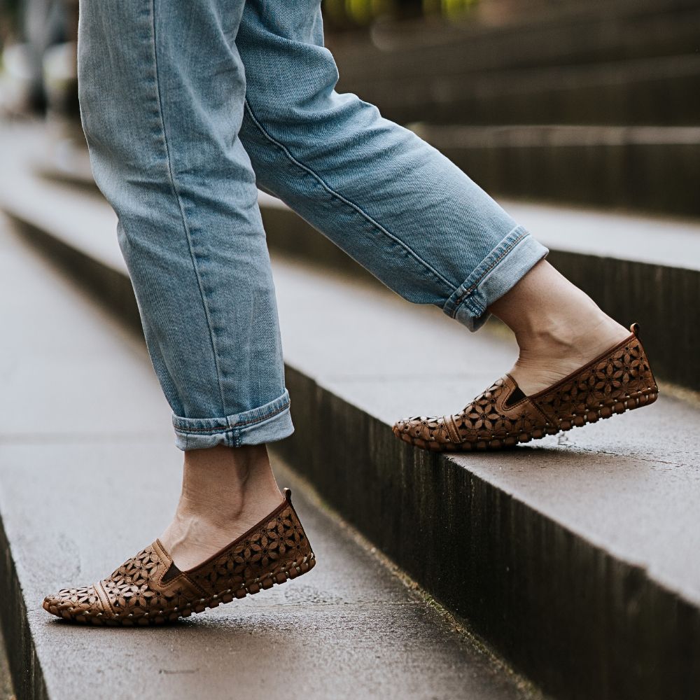 A person wearing blue jeans is walking down some outdoor steps, wearing a pair of brown, laser-cut Turkish leather loafers.