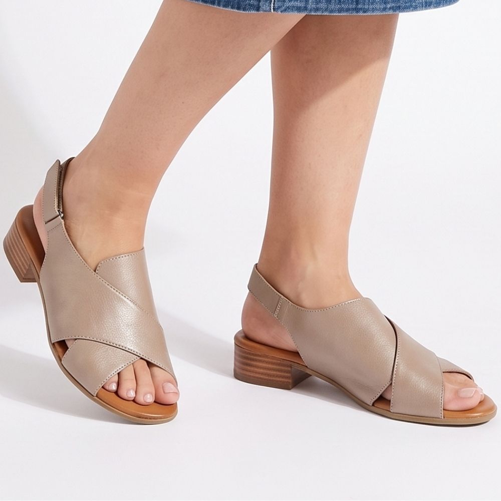 A person wearing taupe crossover leather sandals with a low block heel and rear Velcro strap, standing against a white studio background.