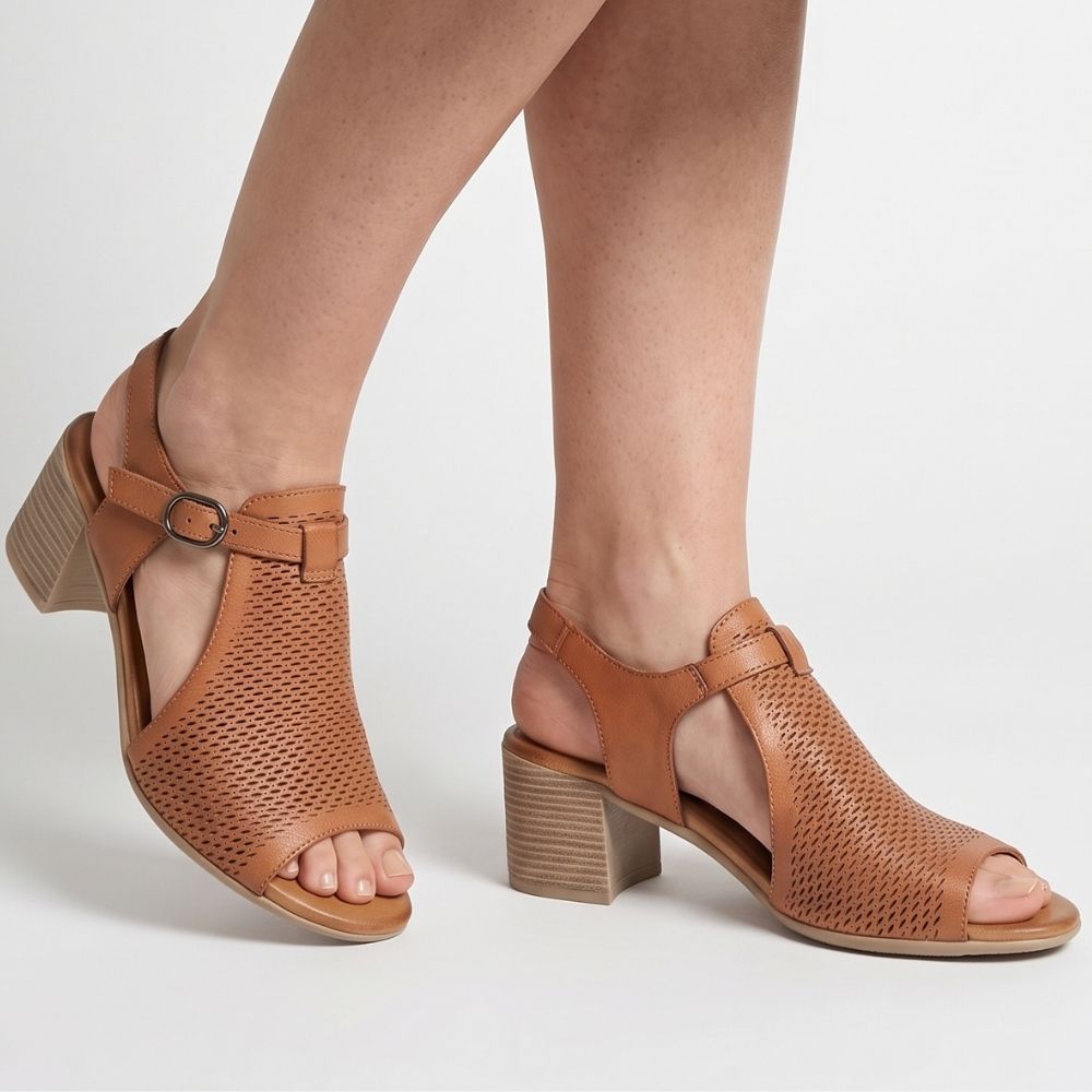 A person wearing tan heeled sandals with a laser-cut leather upper, adjustable bronze buckle and mid-height block heel, against a white background.