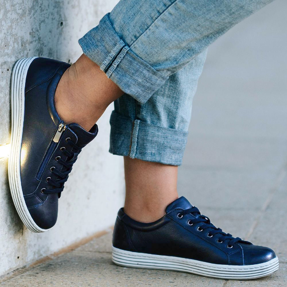 Navy blue sneakers with white soles worn by a person against a light gray wall.