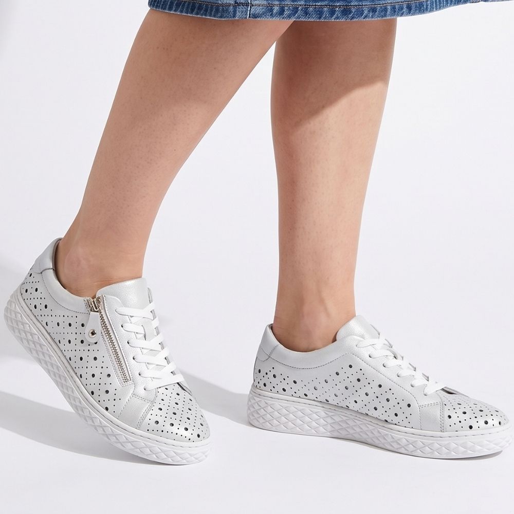 White leather low-top sneakers with perforated detailing, white laces, and a side zipper, shown on a model's feet against a white background.