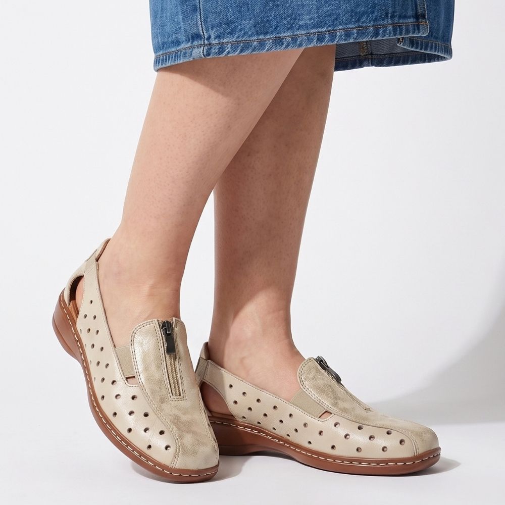 A person wearing a denim skirt and a pair of beige perforated leather slip on shoes with a gold mottle embellished upper, central zip and a brown low wedge outsole, against a studio background.