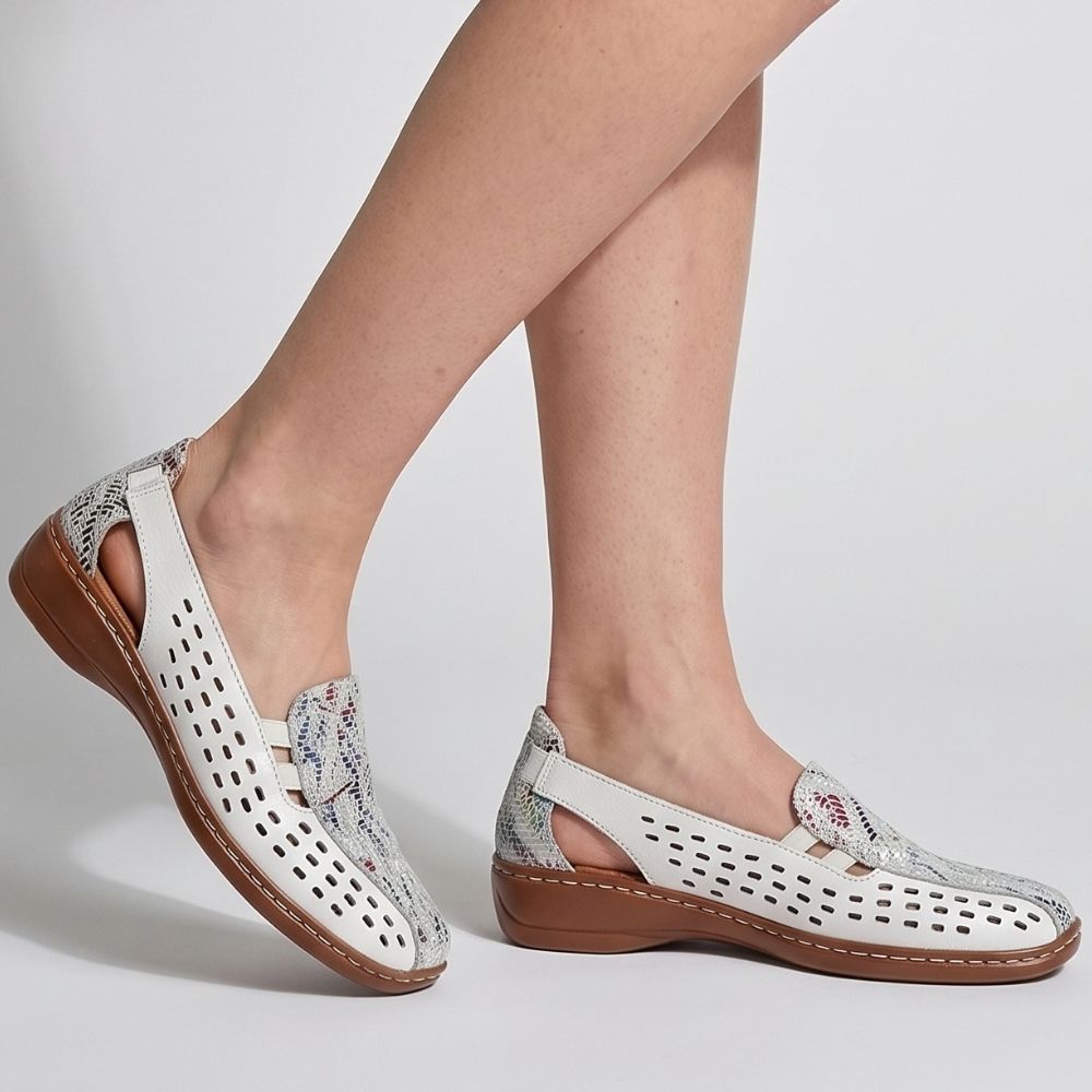 A person wearing a pair of white perforated leather slip on shoes with a multicolour tongue and heel collar and a brown low wedge outsole, against a studio background.