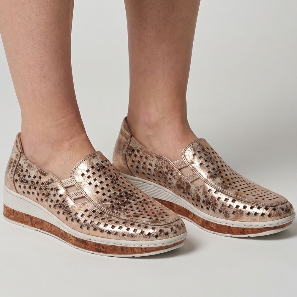 A pair of women's slip-on wedge shoes in a metallic rose gold finish with perforated detailing, worn on a model's feet against a light gray studio. background.