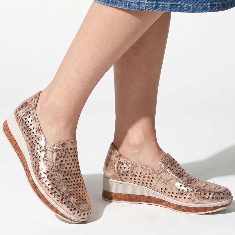 A pair of women's slip-on wedge shoes in a metallic rose gold finish with perforated details on the upper and a cork-textured sole, shown on a model's feet against a white background.
