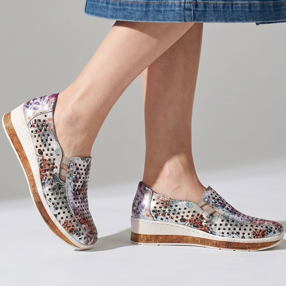 A pair of women's slip-on wedge shoes with a colorful floral print and perforated upper, shown on a model's feet against a light gray studio background.