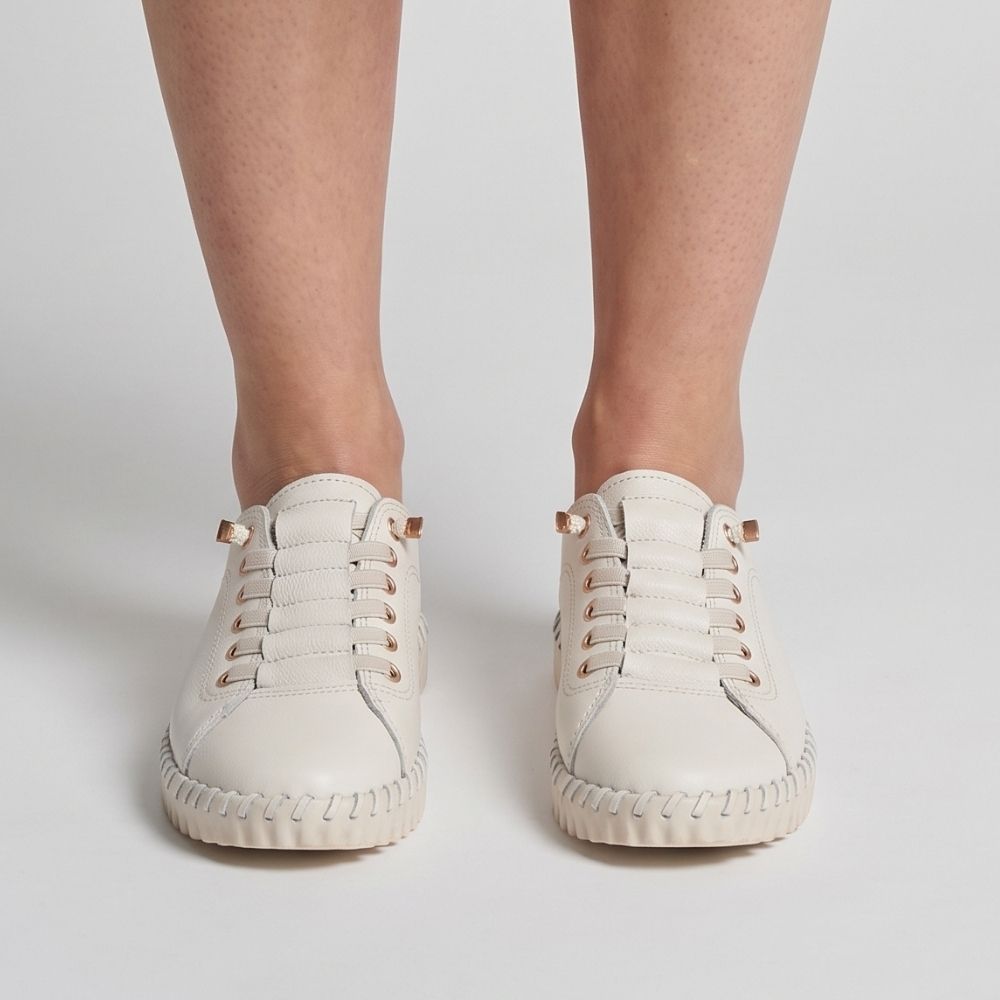 A pair of off-white leather low-top sneakers with gold-tone hardware and leather concealed elastic laces, shown on a model's feet against a light gray background.