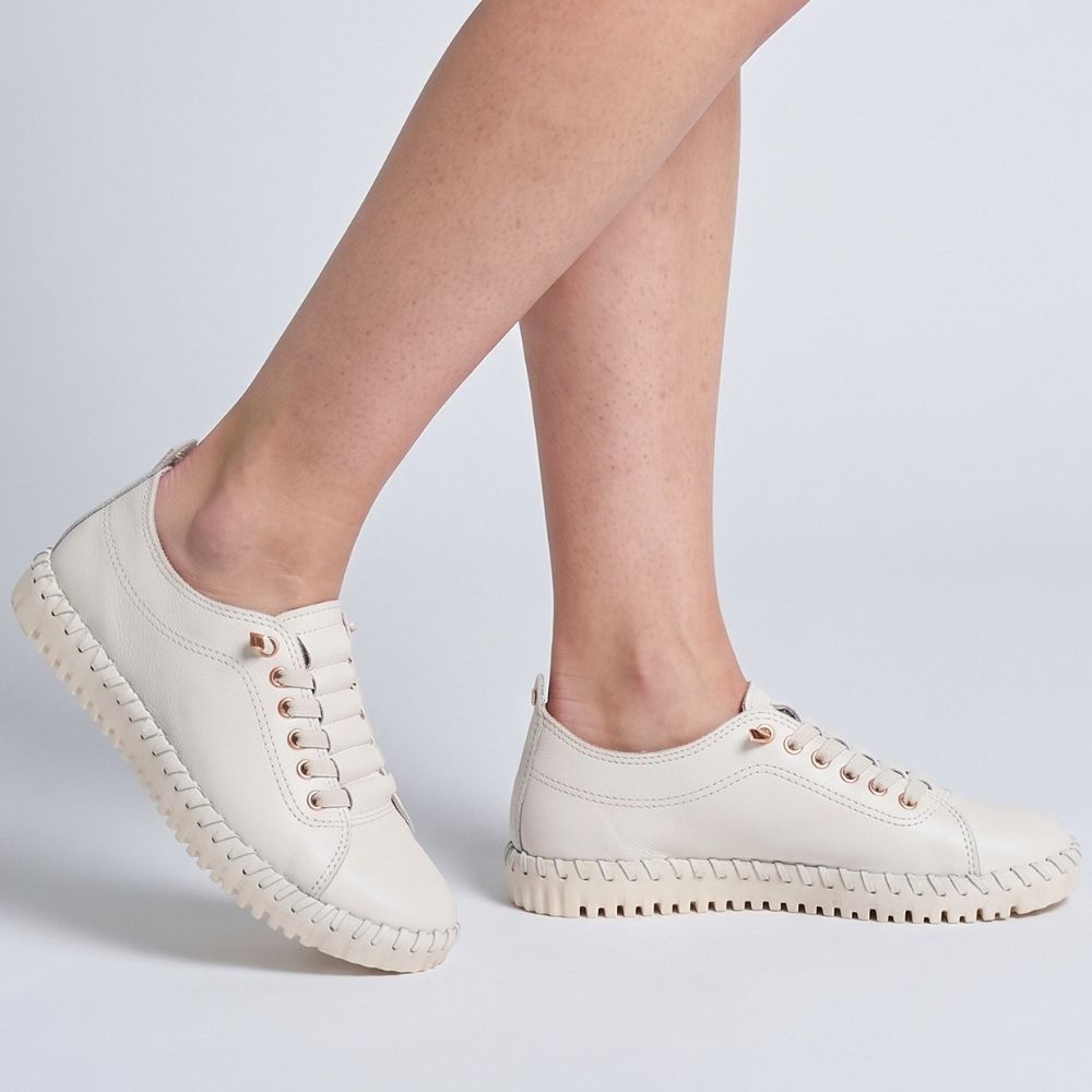 A pair of off-white leather low-top sneakers with concealed laces and a textured, wavy sole, shown on a model's feet against a light gray studio background.