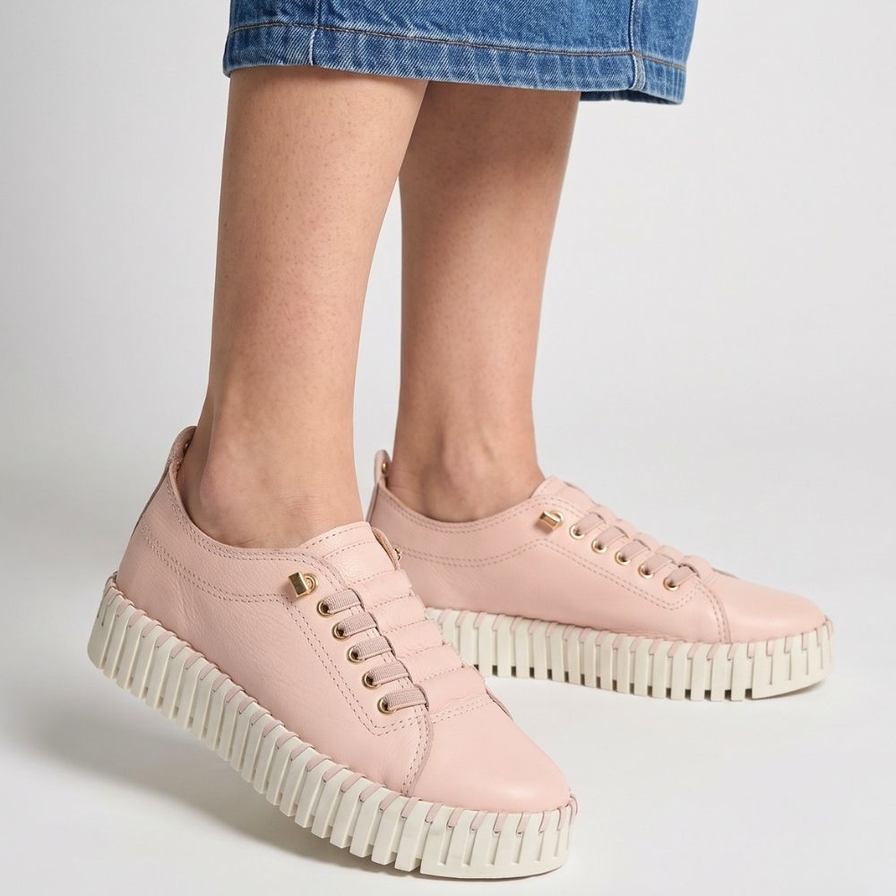 A pair of women's low-top platform sneakers in light pink leather with gold-tone eyelets and ribbed off-white platform soles, shown on a model wearing a blue denim skirt against a light gray studio background.