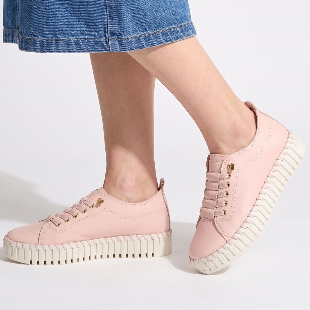 A person wearing light pink low-top platform sneakers with white ridged soles and gold-tone eyelets, shown against a clean white studio background. The model's legs and blue denim skirt are visible.