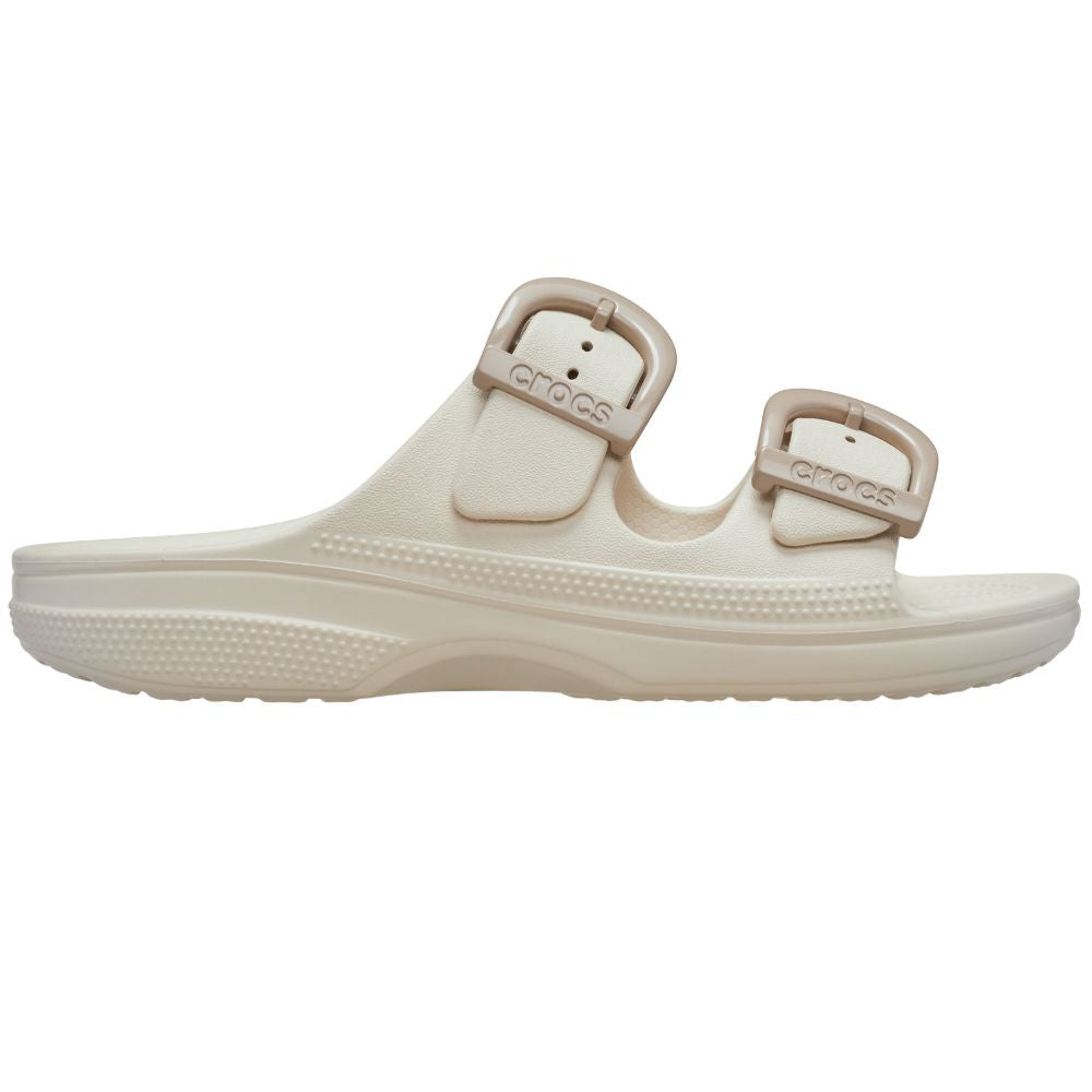 A single Crocs slide sandal in a warm off-white/cream color, featuring dual adjustable straps with taupe enamel buckles and the Crocs logo embossed on them, set against a plain white background.