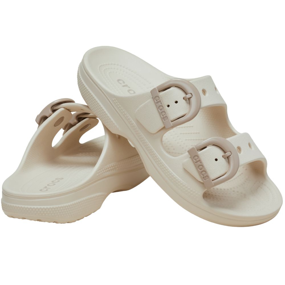 A pair of Crocs double-strap slide sandals in a light cream/off-white color, featuring adjustable taupe enamel buckle straps with the brand name 'crocs' embossed on the buckles.