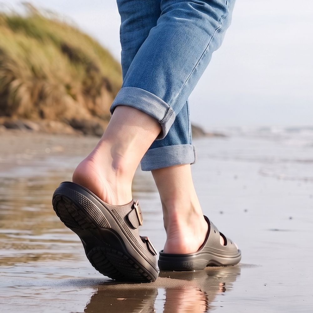A person walking on a beach wearing black Crocs sandals with double straps and light blue rolled-up jeans.