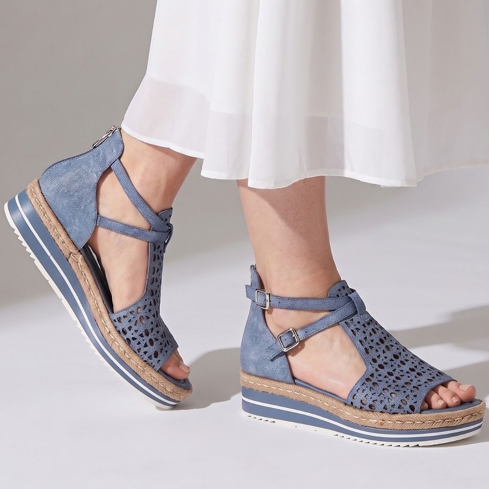 A pair of women's platform wedge sandals in a denim blue leather with laser-cut upper details, ankle straps, and a jute-wrapped sole, shown on a model wearing a white skirt against a light gray studio background.