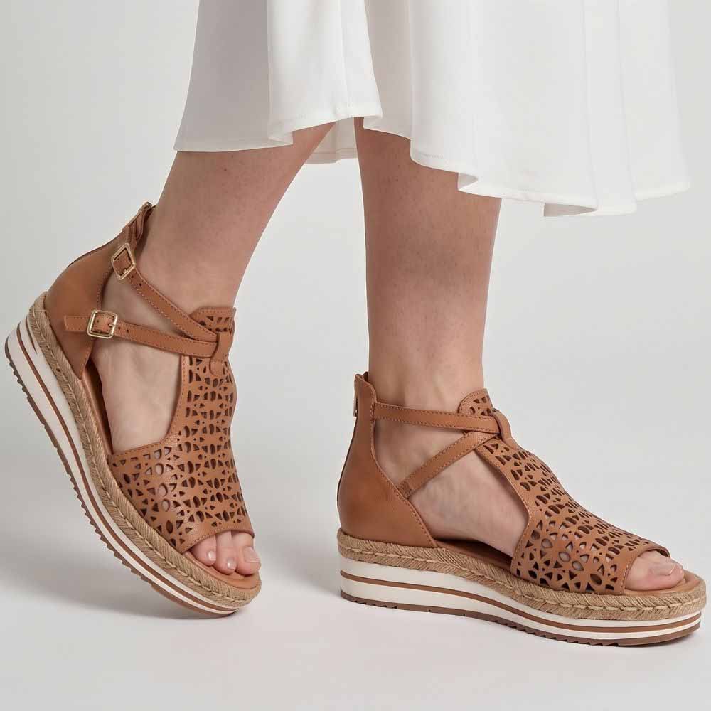 A person wearing a white skirt wearing a pair of tan leather wedge sandals with laser-cut geometric patterns on the toe and upper straps, featuring adjustable ankle straps with gold-tone buckles and a braided jute-wrapped wedge platform sole, against a grey studio background.