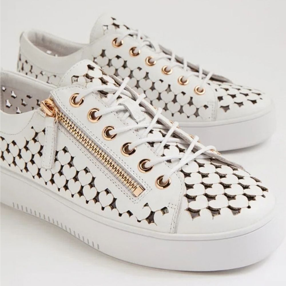 White low-top leather sneakers with a heart shaped cutout pattern, white laces, a gold-tone zipper, and gold-tone eyelets, shown on a light gray background.