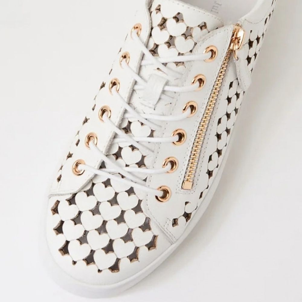 White leather sneaker featuring a heart-shaped cutout pattern, white laces, gold-tone eyelets, and a gold zipper on the side.