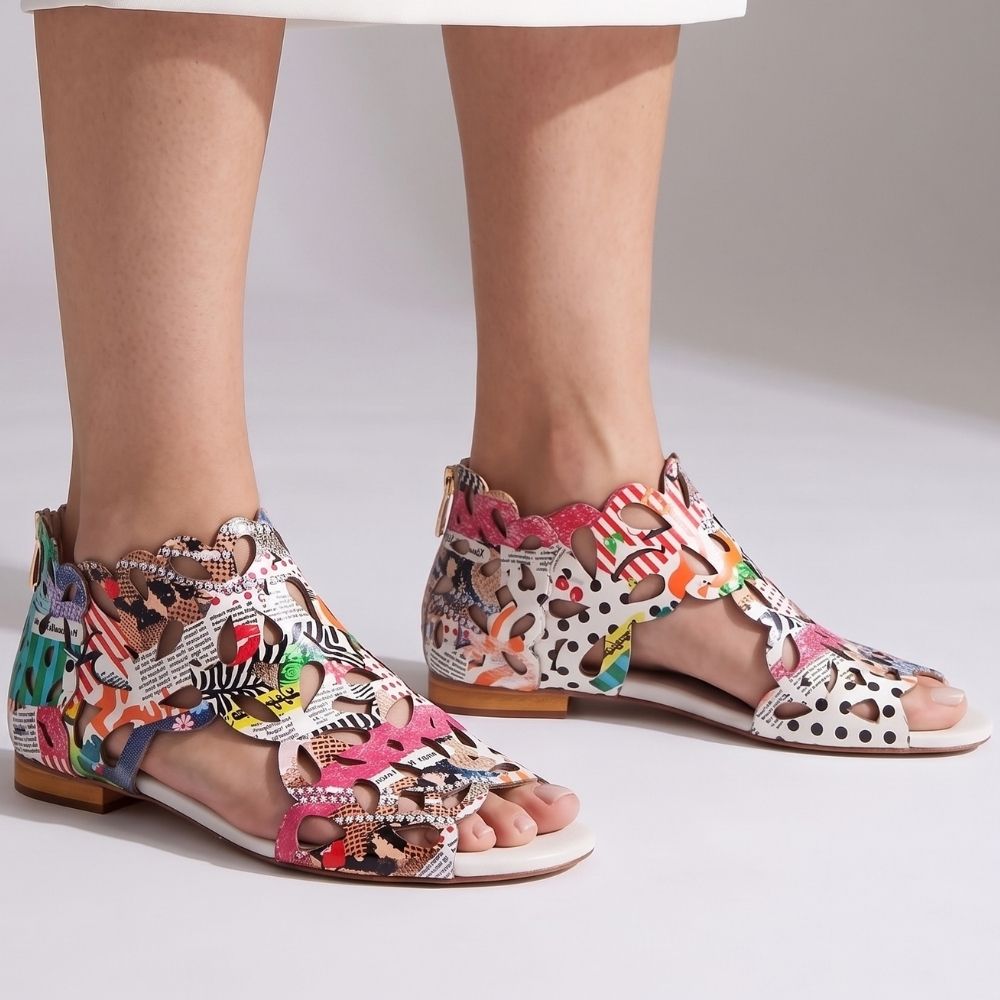 A pair of women's flat sandals with an intricate multicolor abstract print, shown on a model's feet against a light gray studio background.