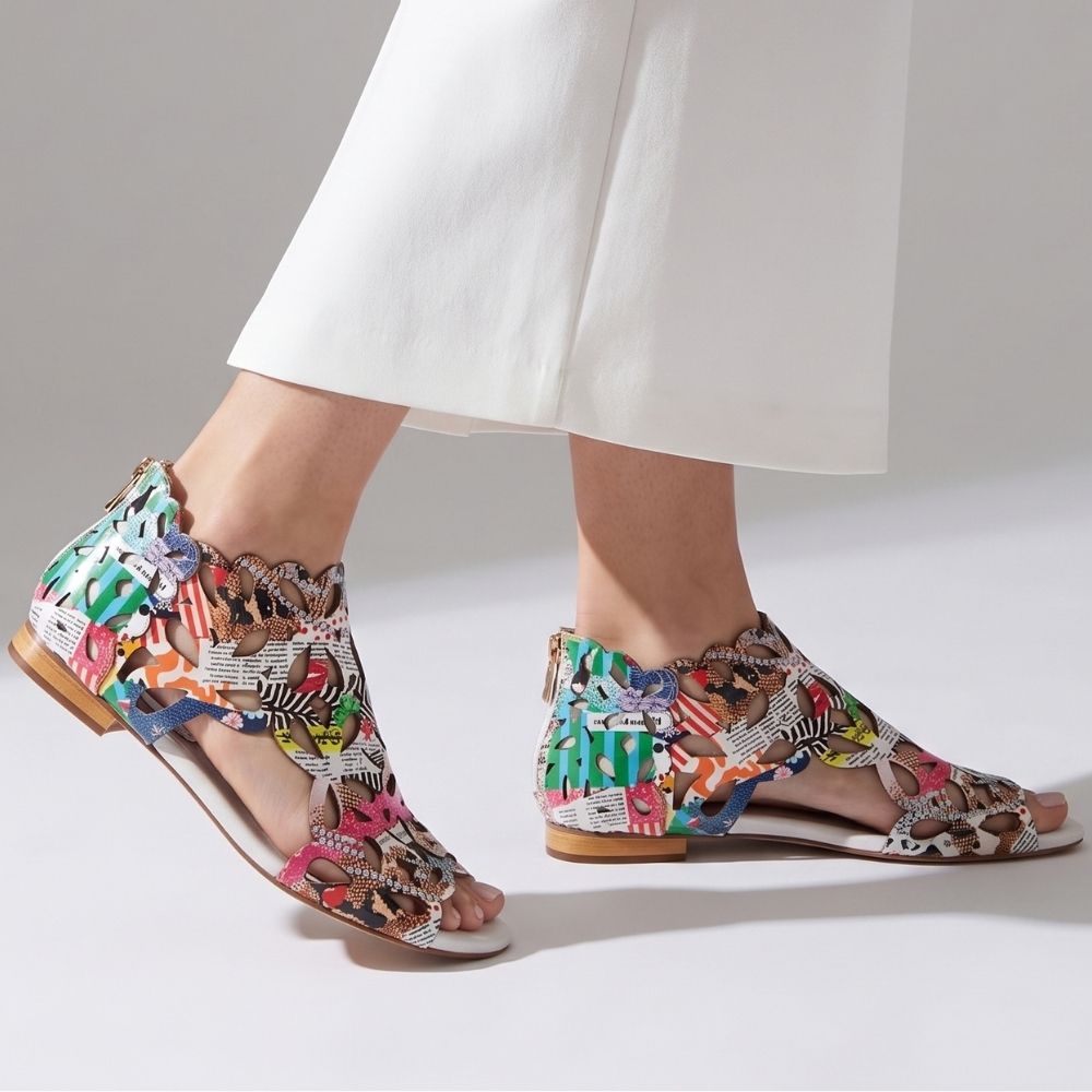 A pair of women's flat sandals with a colorful, multicolored abstract patterned upper and a cream sole, shown on a model wearing white pants against a light gray studio background.