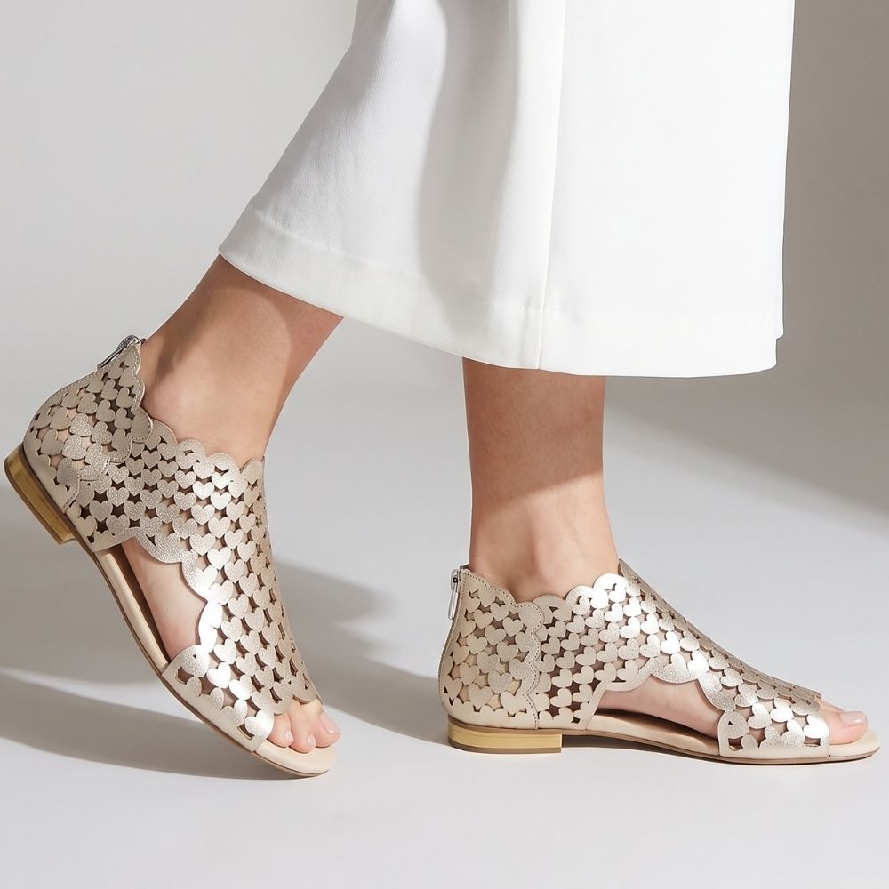 A pair of metallic gold cutout flat sandals worn with a white skirt, shown from the side against a neutral light gray background.