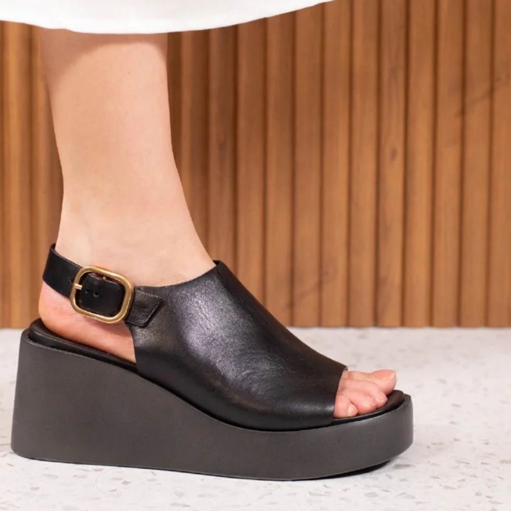 A person wearing a black leather wedge sandal with an ankle strap and gold-tone buckle, shown on a foot against a wood-paneled background.