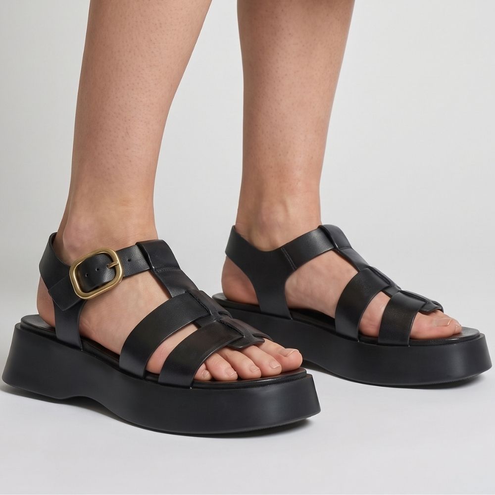 Black leather platform sandals with wide straps and a gold buckle, shown on a model's feet against a light gray background.