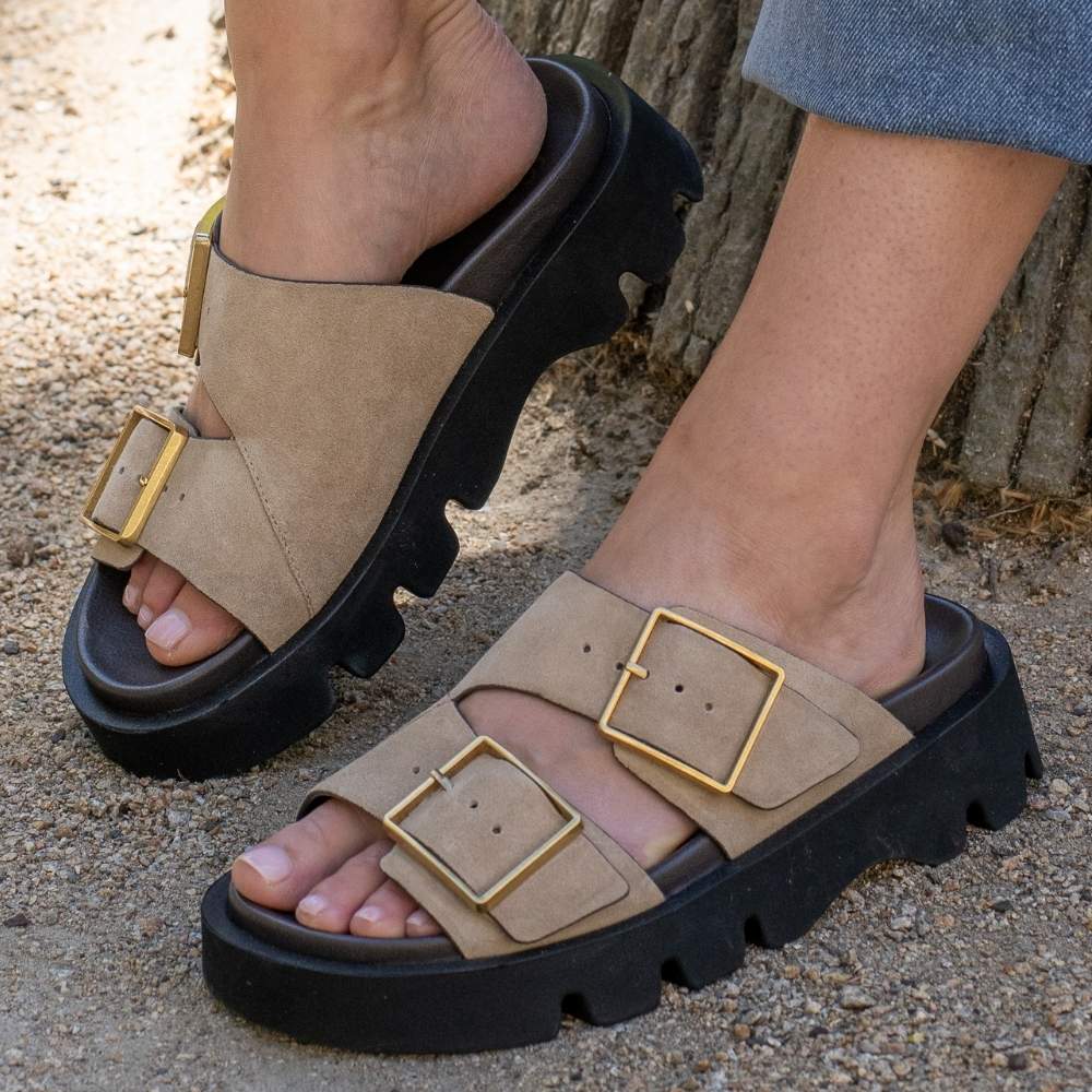 A close-up of a person wearing chunky platform slide sandals with wide beige suede leather straps, gold-tone buckle details, and thick black lug soles, standing outdoors on gravel near a tree trunk.