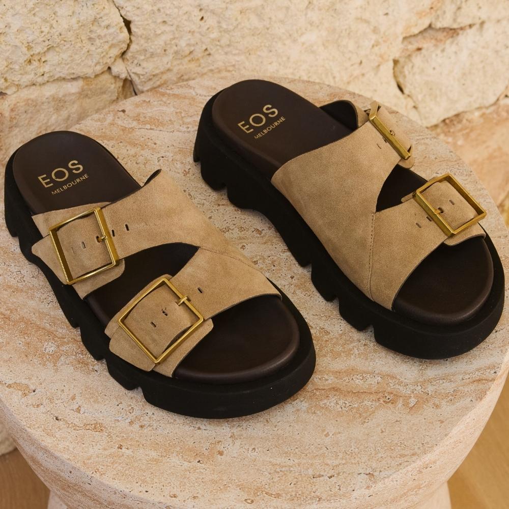 A pair of chunky platform slide sandals in beige suede with gold-tone buckle straps and a black sole, displayed on a round stone pedestal against a stone wall background.