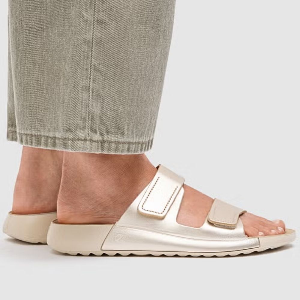 Person standing up wearing grey jeans and pale gold slip on leather sandals with two adjustable straps on a white background