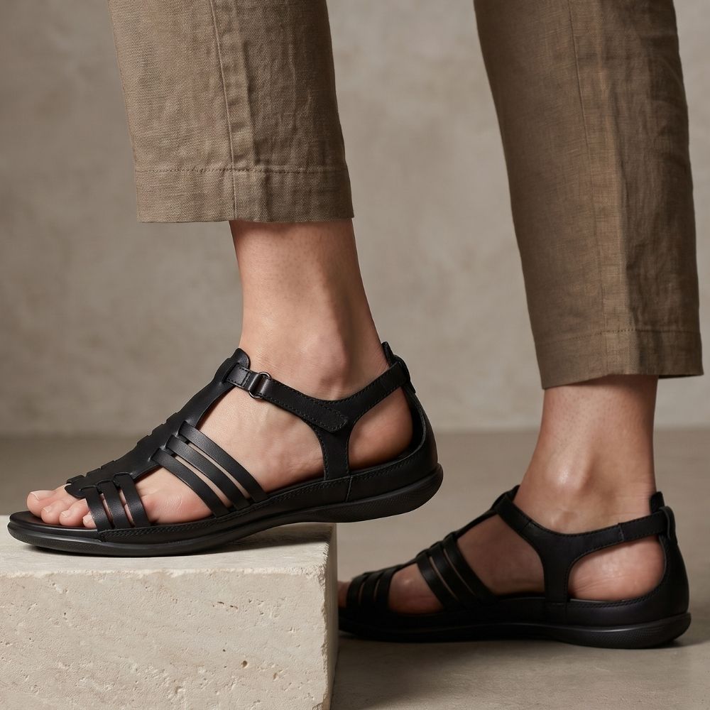 A pair of black leather strappy flat sandals worn with brown linen pants, shown on a model's feet against a neutral textured beige background.