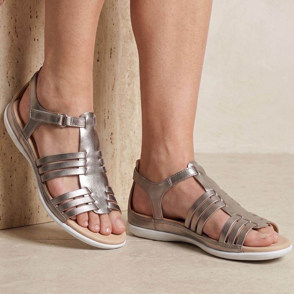 Metallic pale gold leather strappy sandals worn on a model's feet, shown against a neutral beige textured background.