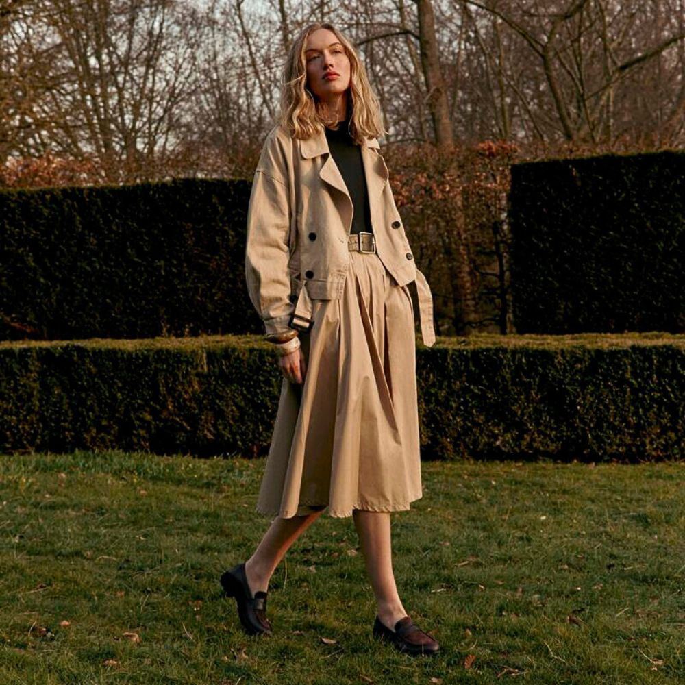 Woman in a beige trench coat standing in a garden with hedges and trees in the background.