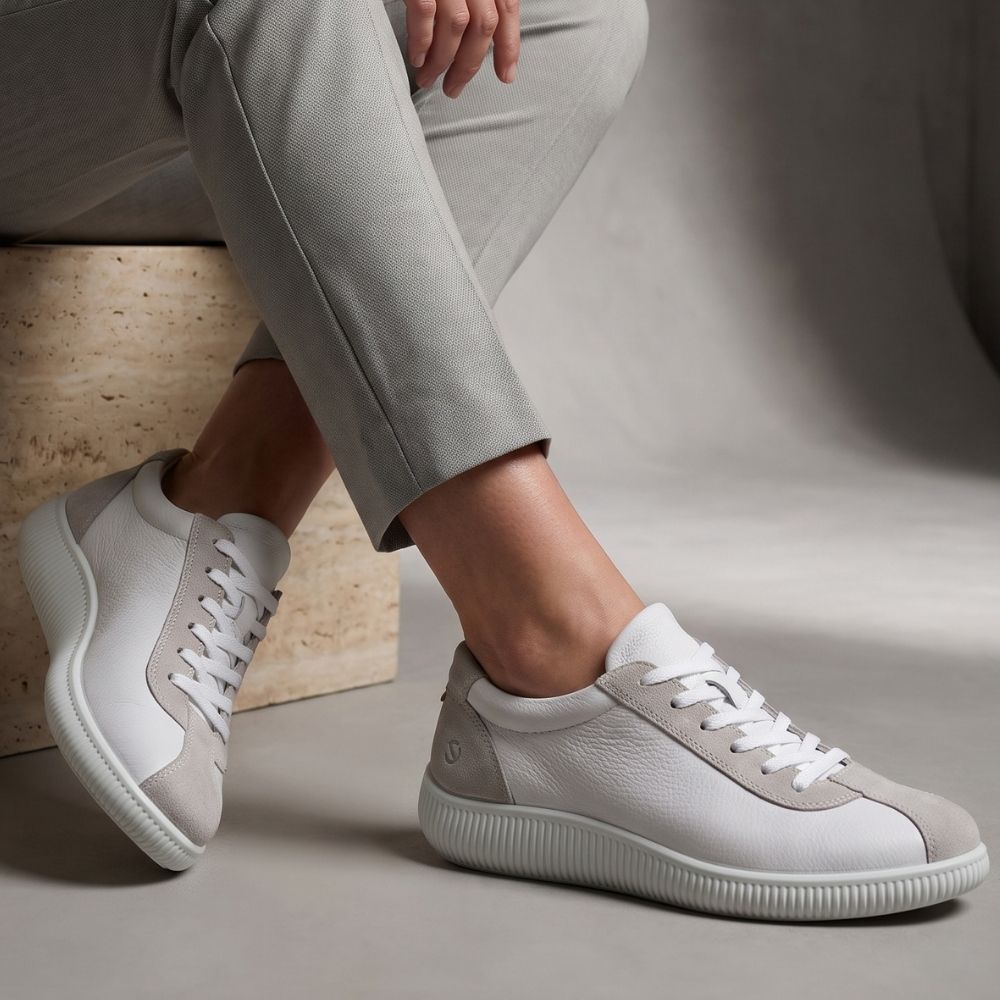 A pair of low-top white leather sneakers with light gray suede overlays, shown on a model wearing light gray trousers. The model is seated on a textured stone block against a neutral gray studio background.