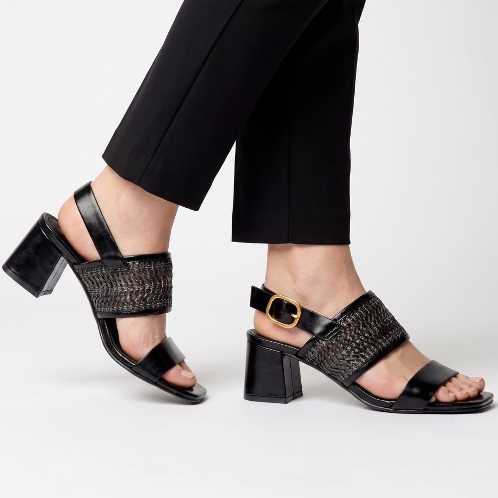 A pair of women's black block-heel sandals with textured woven straps and a gold buckle, shown on a model wearing black trousers against a light gray studio background.