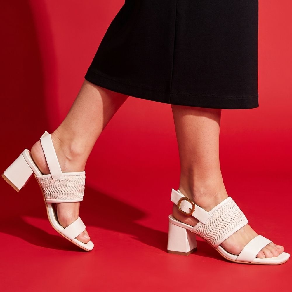 A person wearing a long black skirt with white block heeled sandals with woven upper straps and gold buckles, against a red background.