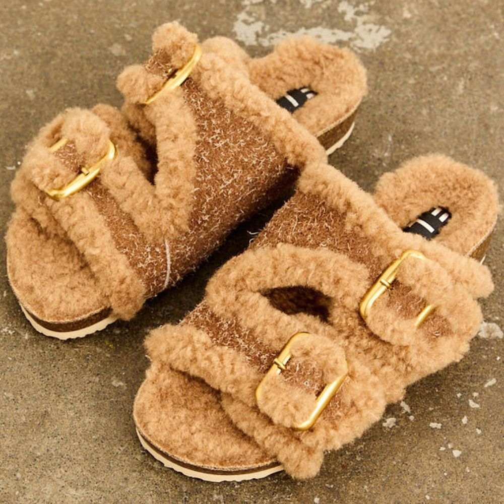 Pair of caramel leather slip-ons with fluffy lining and gold buckles on a textured surface