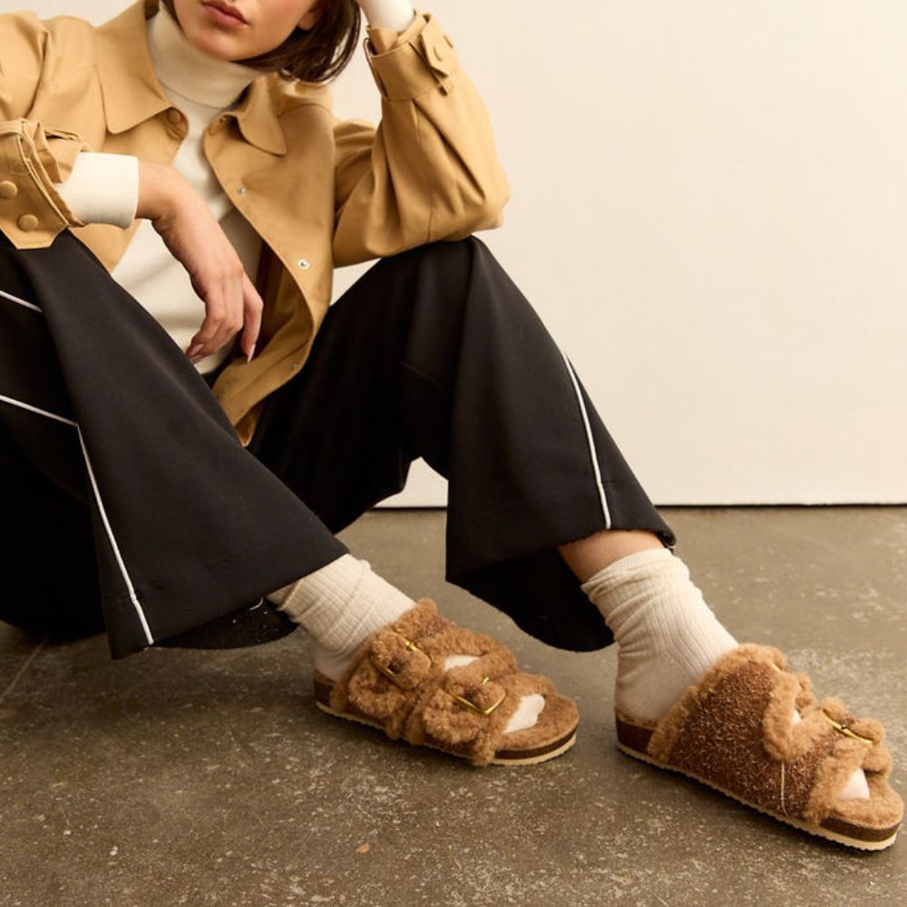 Person wearing caramel leather slip-ons with fluffy lining and gold buckles on a textured surface