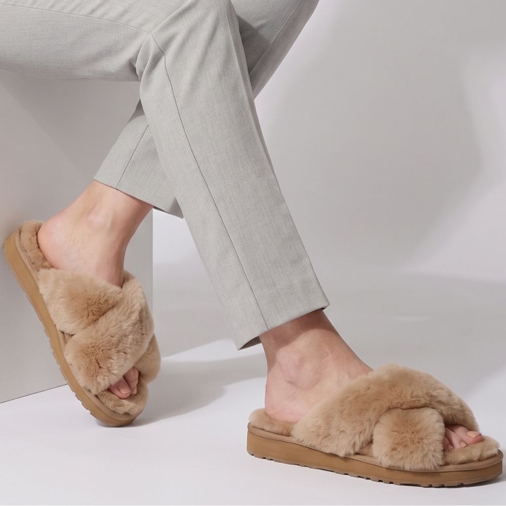 A person wearing sheepskin slippers with thick tan soles, paired with light gray trousers, seated on a white block against a white studio background.