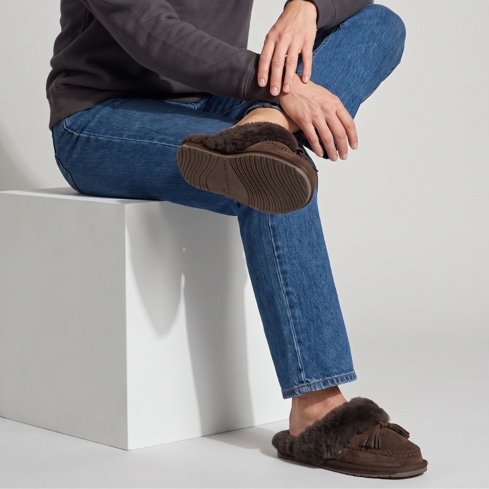 A lady with blue jeans sitting on a white stool cross-legged, wearing dark brown suede slip-on slippers with a thick sheepskin lining on a white background.