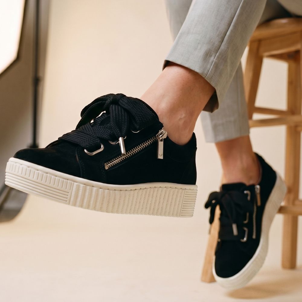 Person sitting on a stool wearing grey pants and black suede sneakers with a platform sole, side zip and statement laces