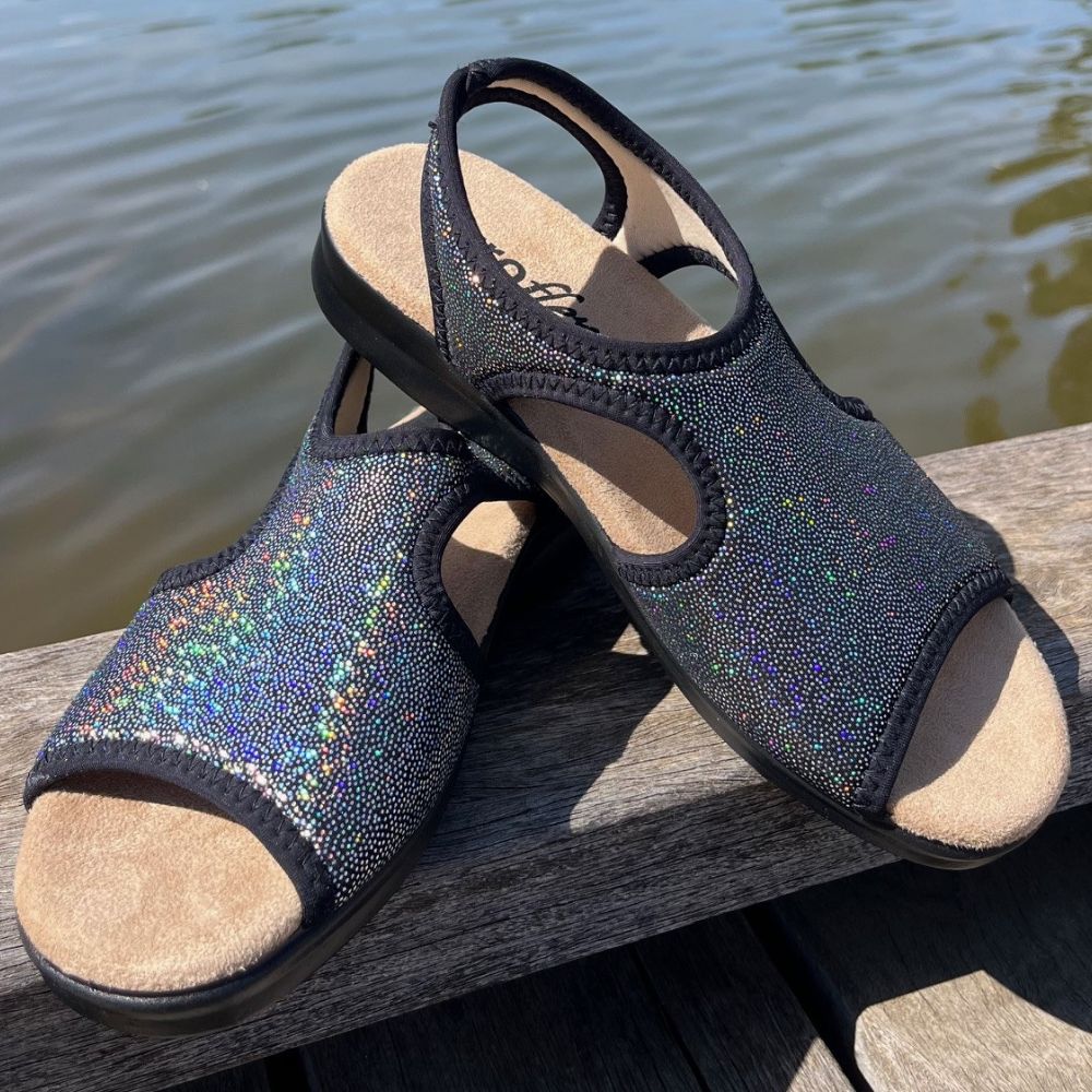 Pair of black sandals with glittery texture on a wooden surface by water.