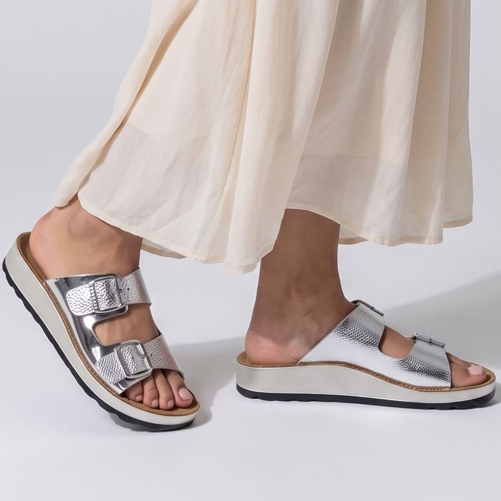 A person wearing a floaty skirt with silver leather slide sandals with two adjustable buckle straps and a lightweight sole, on a white studio background.