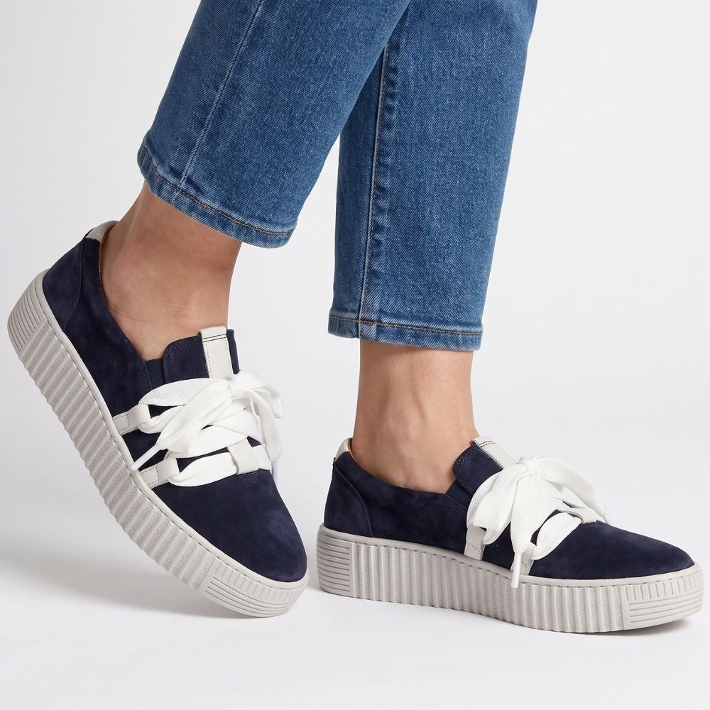 Person wearing blue jeans with navy suede sneakers with a white cleated sole and decorative white laces on a white background.