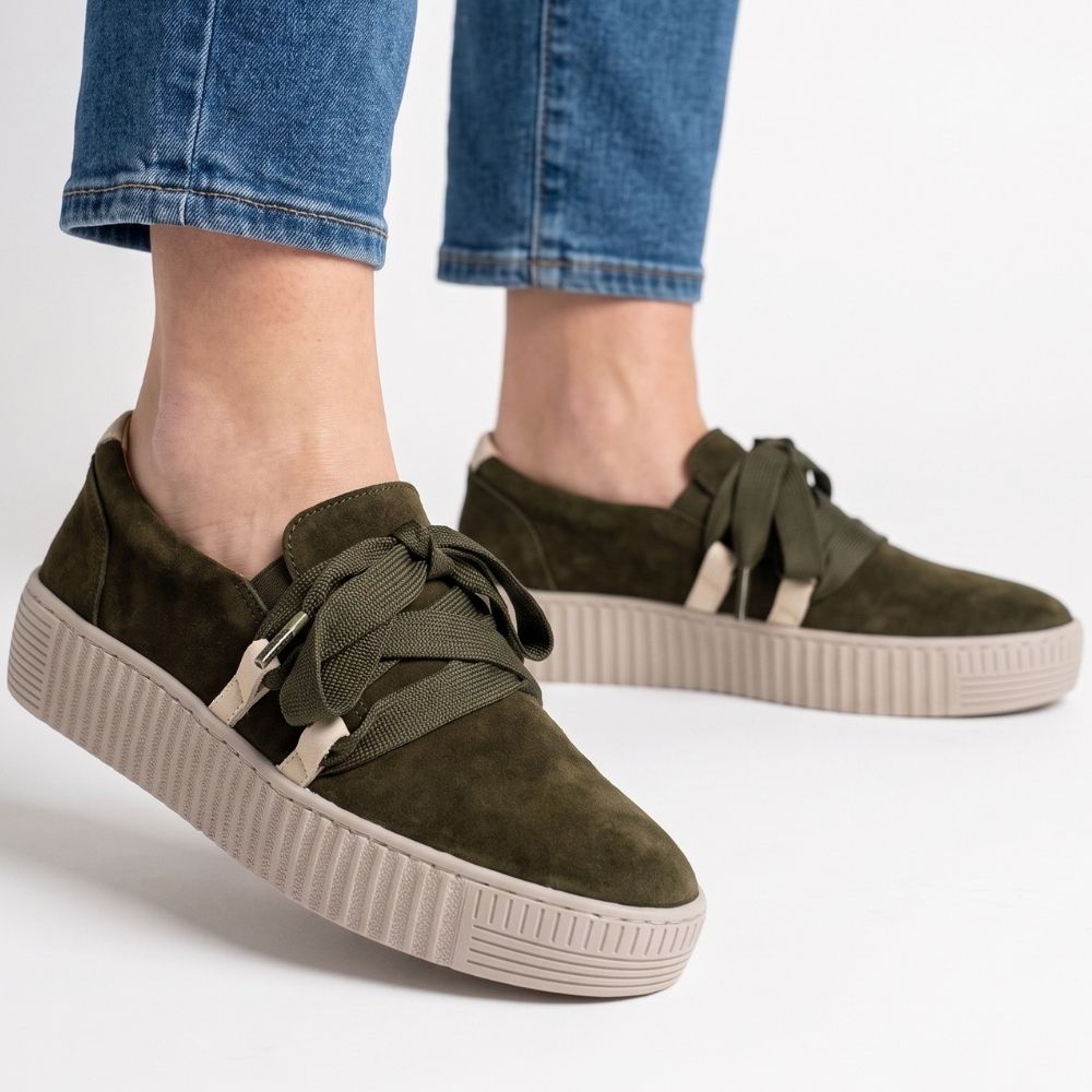 Person wearing blue cropped jeans and olive suede leather sneakers with decorative laces, on a white background.