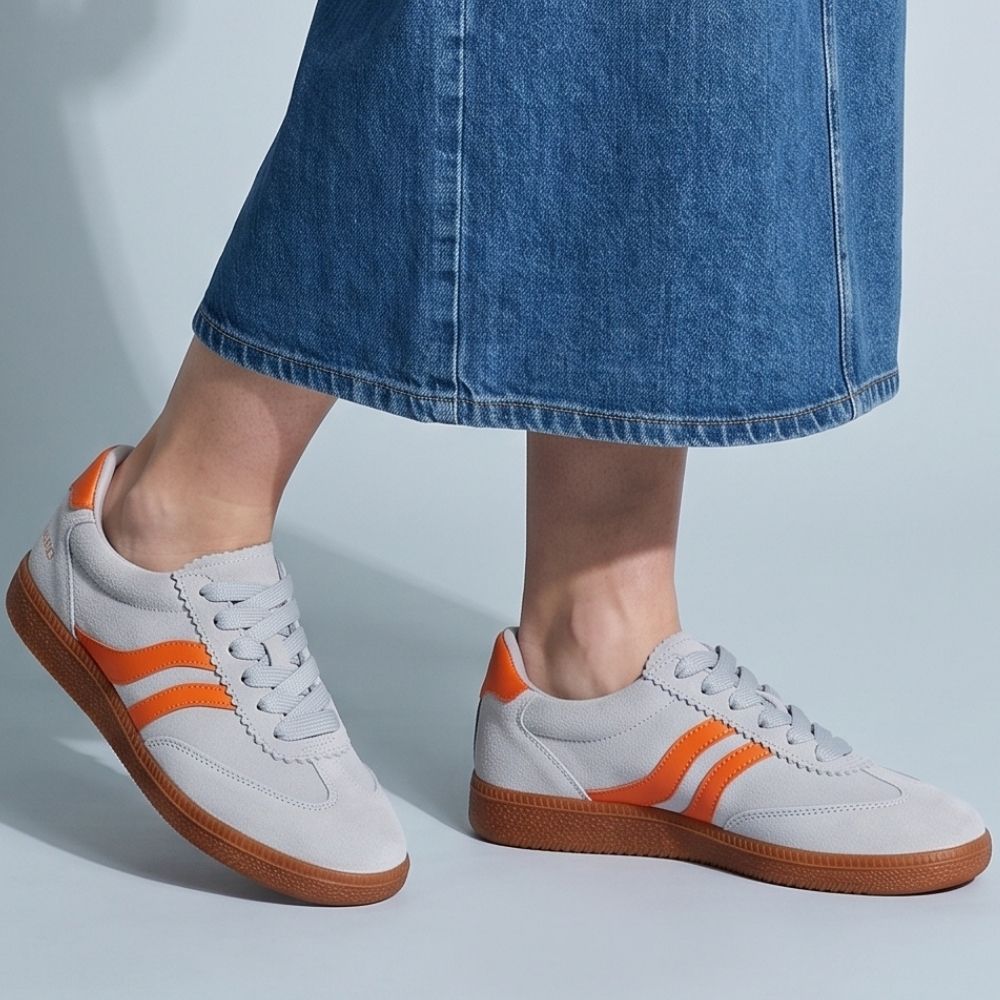 Person wearing a long denim skirt and pale blue suede lace-up sneakers with orange details and a brown rubber sole, against a blue studio background.