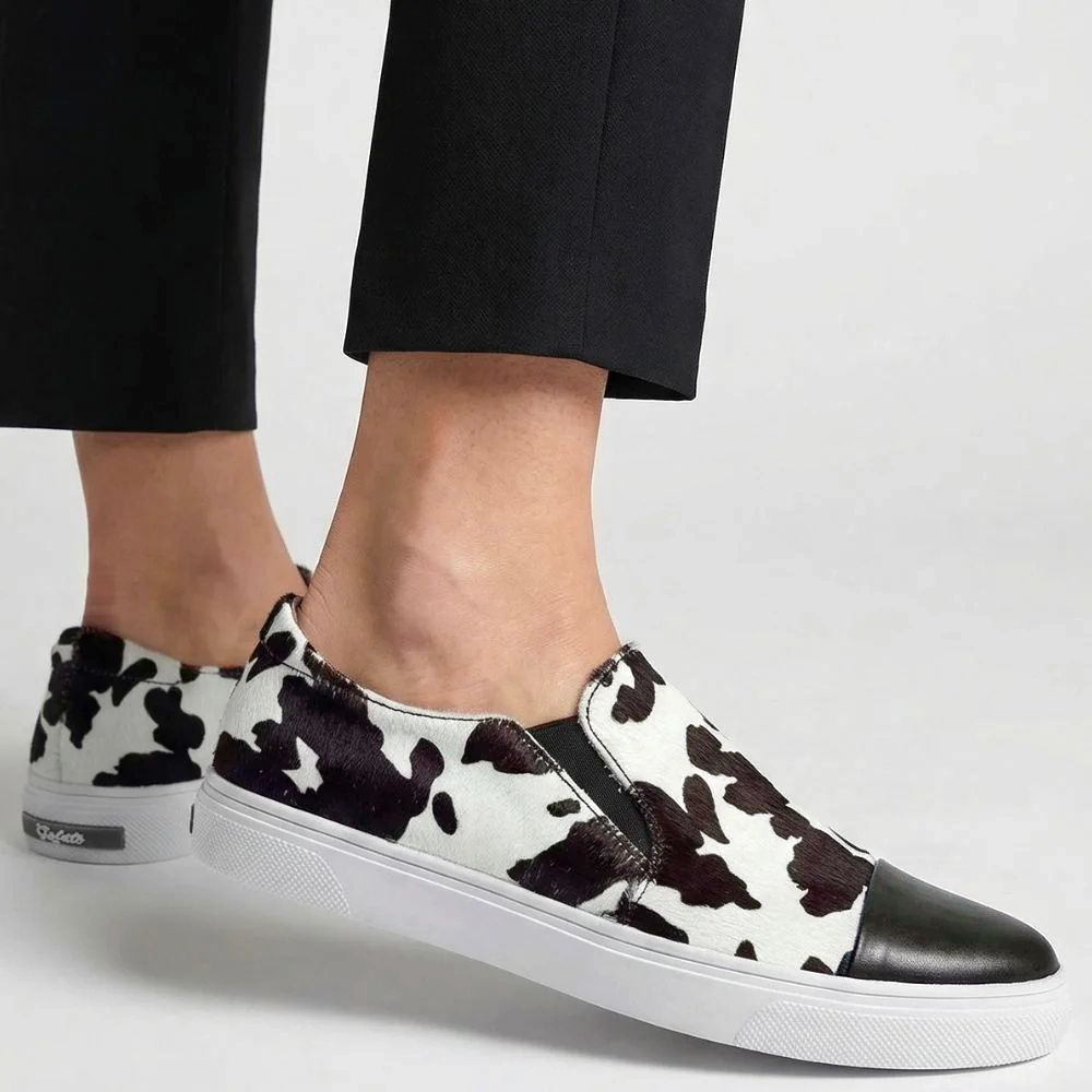 Brown and white cow print slip on loafer sneakers worn with black pants on a light gray background